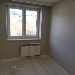 Artluk - Świeżo pomalowane pomieszczenie z białymi panelami podłogowymi, grzejnikiem pod oknem i widokiem na drzewa za oknem. Na podłodze widoczny ręcznik papierowy i torba narzędziowa.