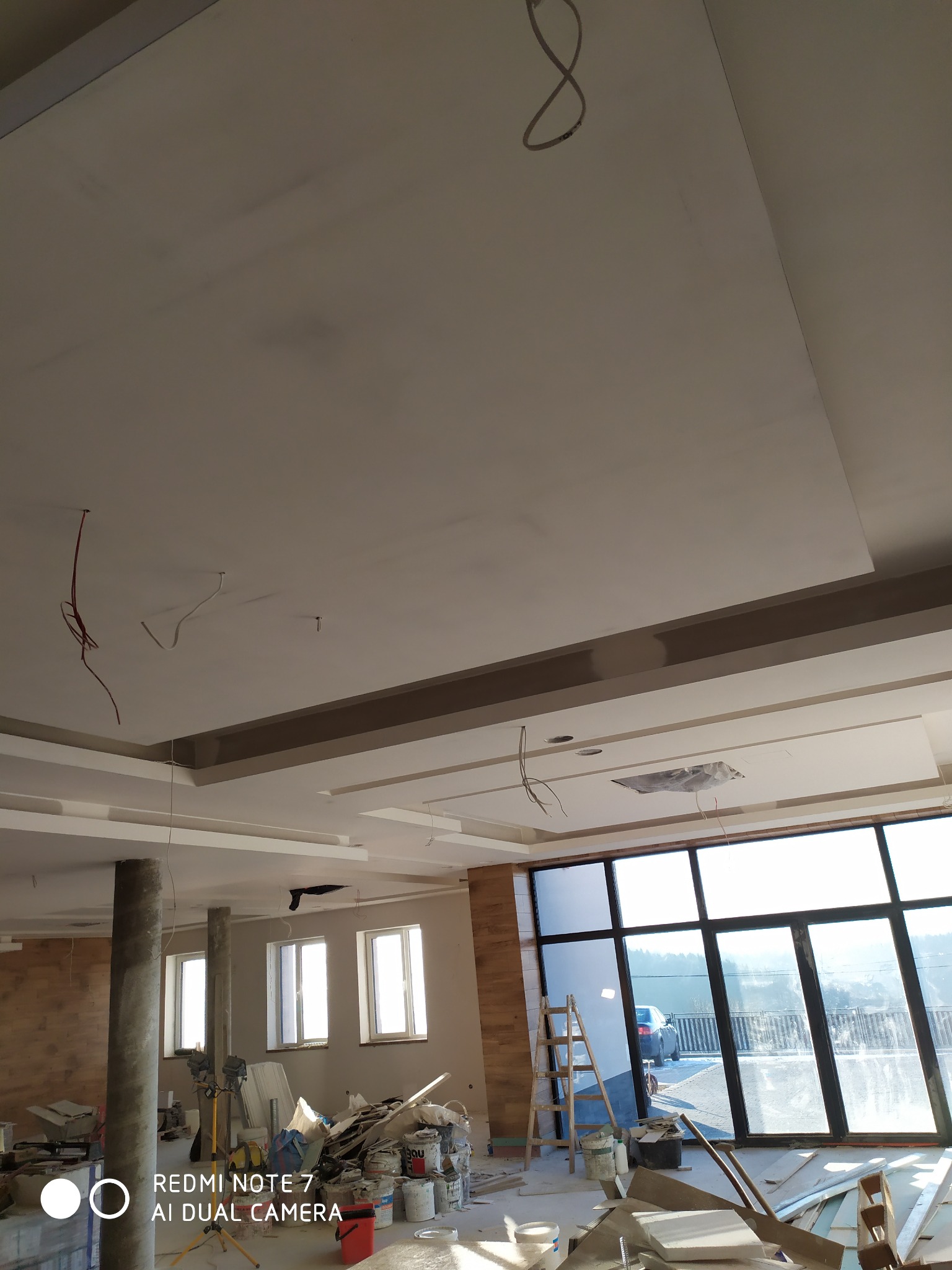 Wnętrze w trakcie prac wykończeniowych: widoczny sufit podwieszany z elementami dekoracyjnymi, wystające przewody elektryczne, nieporządek na podłodze, puszki z farbą i narzędzia.