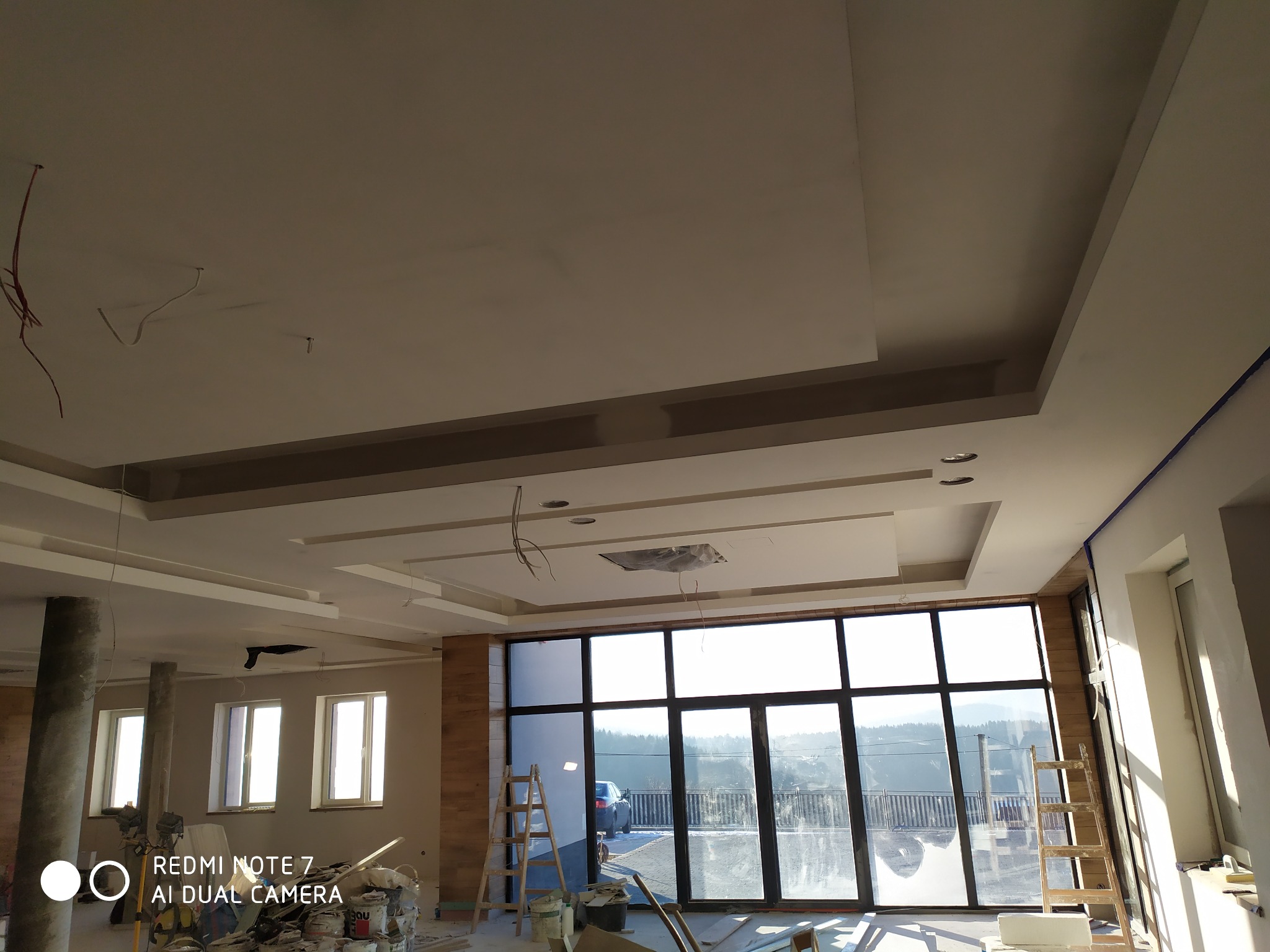 Wnętrze w trakcie prac wykończeniowych: sufit z dekoracyjnymi elementami, widoczne kable, duże okno z widokiem na zewnątrz, drabina, puszki z farbą i narzędzia malarskie.