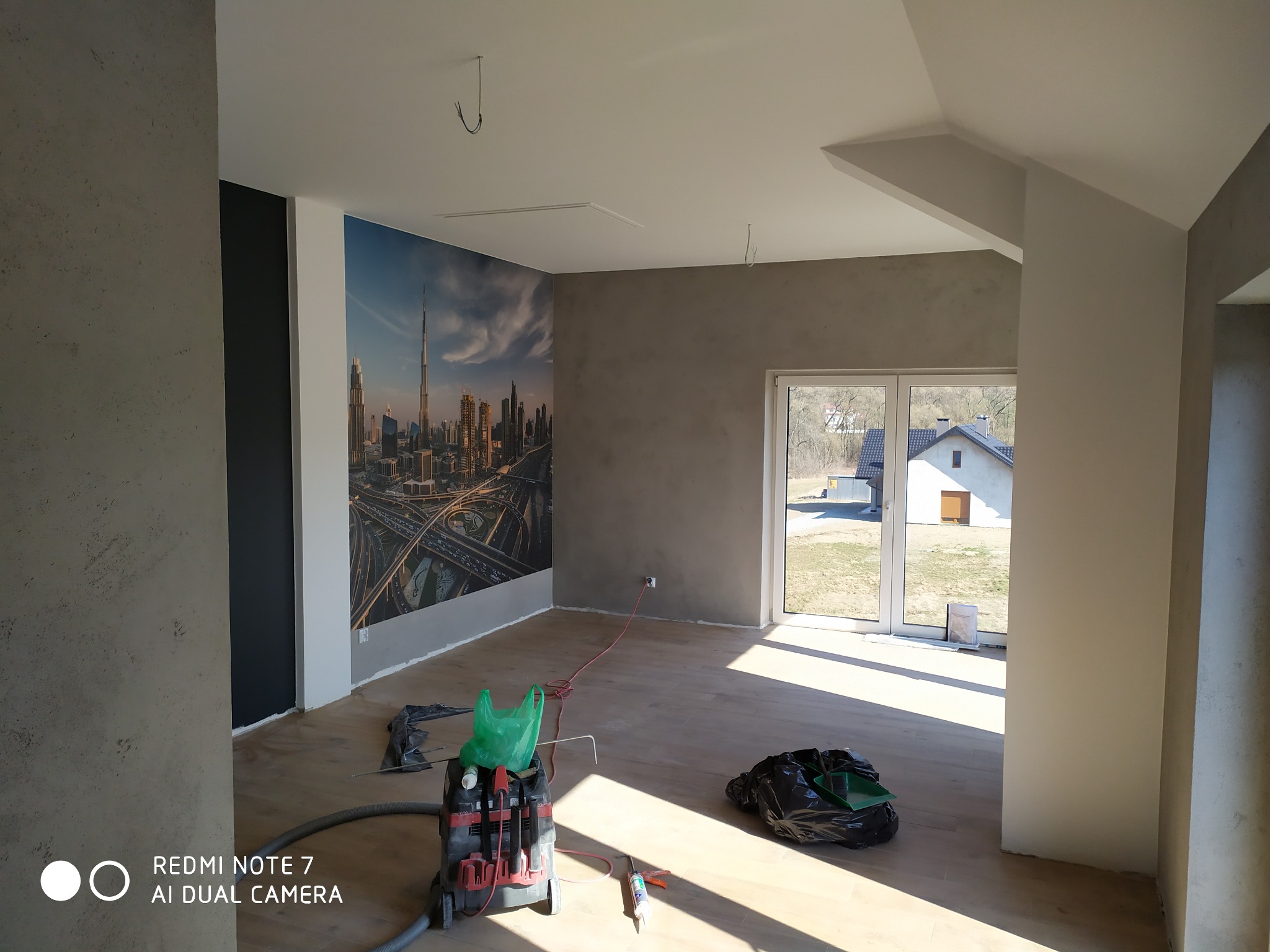 Wnętrze pokoju po pracach wykończeniowych, ściany pokryte szarą strukturą, fototapeta z panoramą Dubaju, widoczny odkurzacz przemysłowy i akcesoria malarskie, jasna podłoga z paneli.