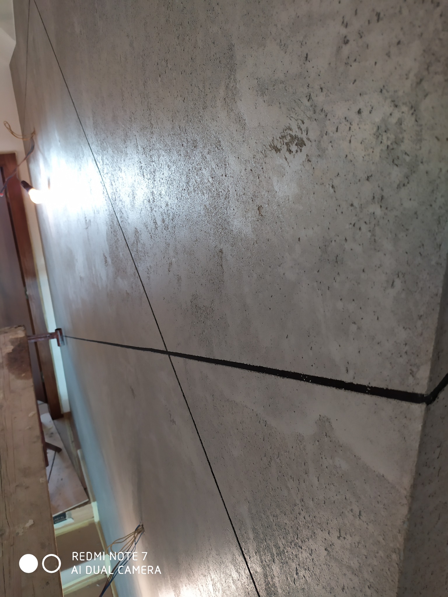 Ściana z surowym wykończeniem, widoczne ślady szpachlowania i linie wyznaczające przyszły podział, oświetlona żarówką na kablu.