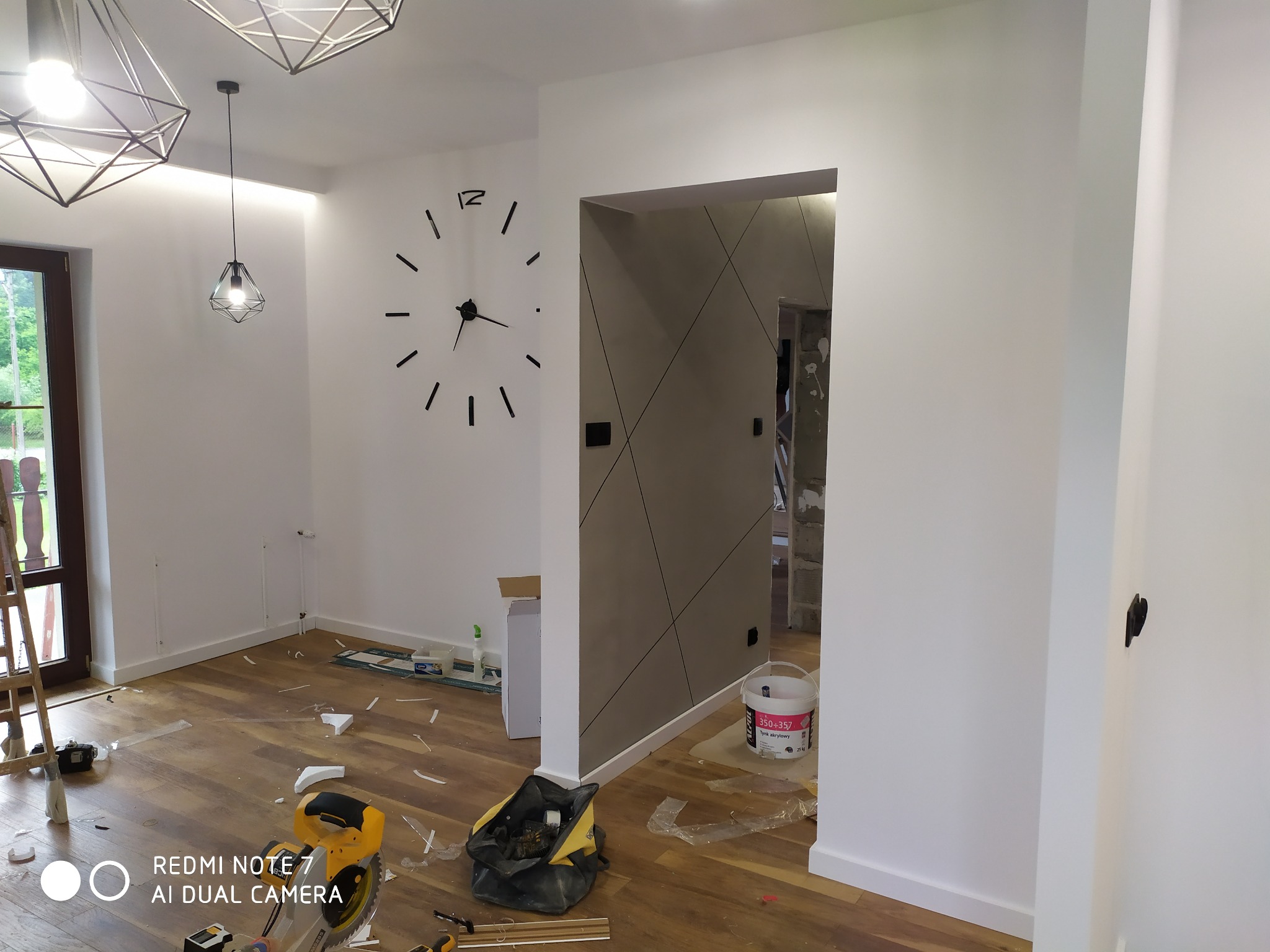 Wnętrze mieszkania w trakcie prac wykończeniowych: świeżo pomalowane białe ściany, drewniana podłoga z widocznymi śladami prac, nowoczesny zegar ścienny, geometryczne lampy sufitowe i widoczny...