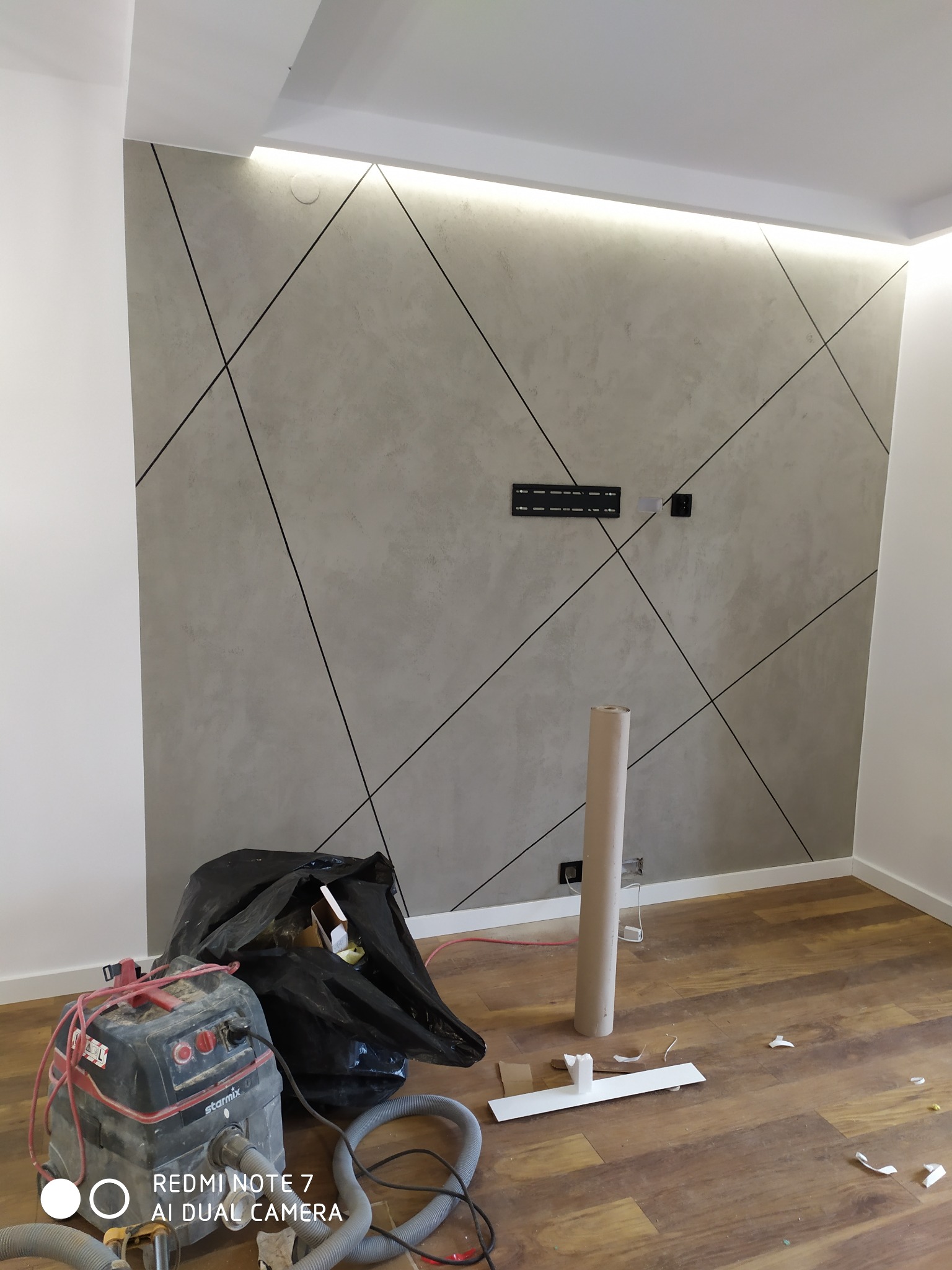 Ściana pokryta dekoracyjną szarą strukturą z czarnymi liniami, podświetlona listwą LED, widoczny odkurzacz przemysłowy, rolka papieru i śmieci na podłodze z paneli.