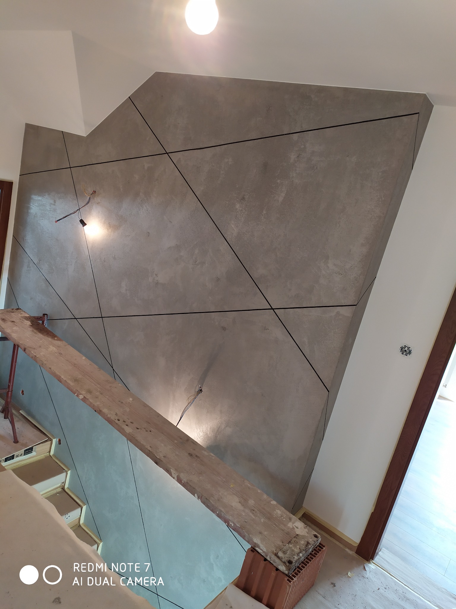 Ściana pokryta szarą dekoracyjną fakturą z czarnymi liniami tworzącymi geometryczny wzór, widoczny fragment rusztowania i niedokończone wykończenie gniazdka elektrycznego.