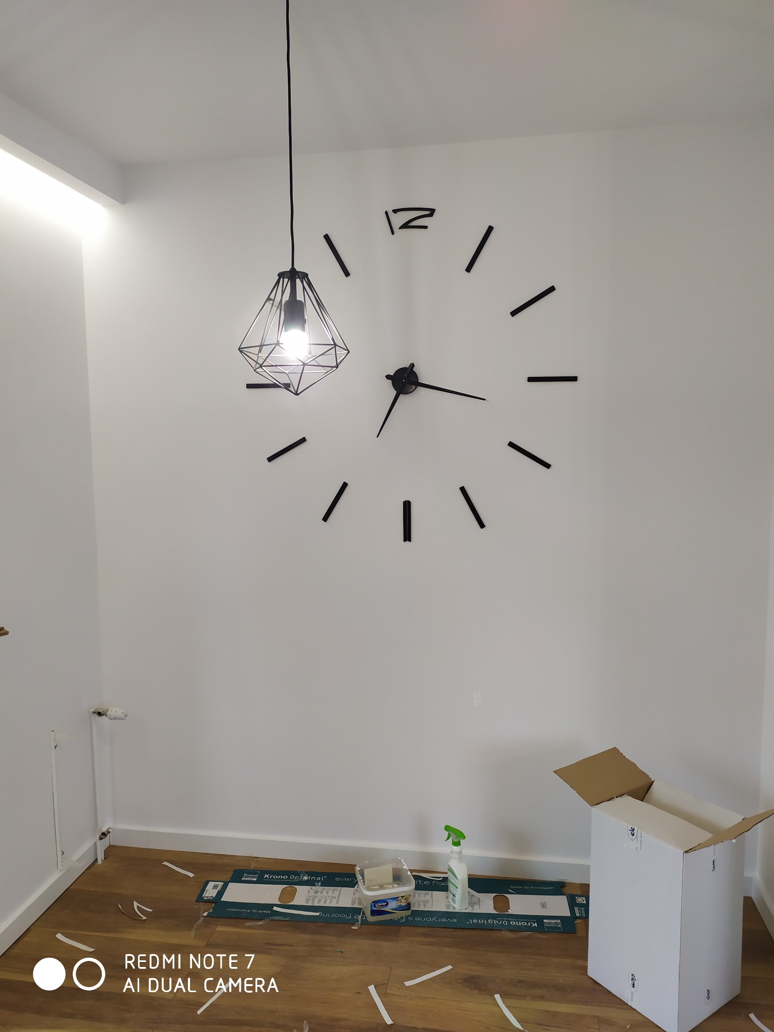 Wnętrze pokoju z dużym, czarnym zegarem naklejonym na białą ścianę, wiszącą metalową lampą w kształcie diamentu, fragmentem podłogi z panelami i kartonem po panelach oraz akcesoriami do montażu...