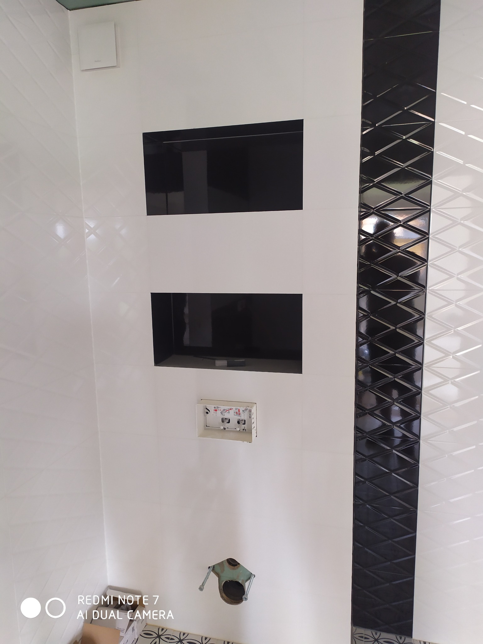 Nowoczesna łazienka z białymi płytkami o delikatnej fakturze, czarnymi wnękami i pionowym pasem dekoracyjnym z czarnych, błyszczących rombów. Widoczny stelaż podtynkowy WC i fragment podłogi...