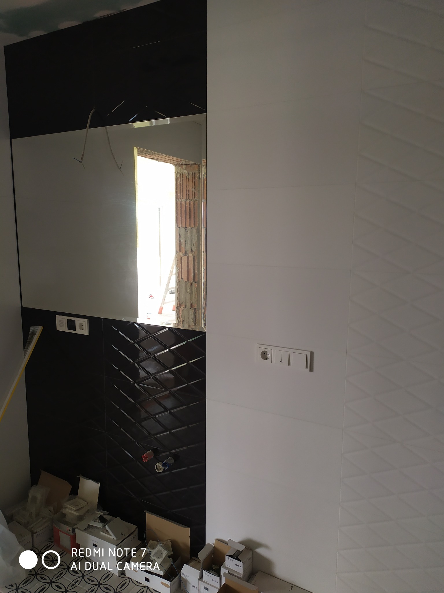 Częściowo wykończona łazienka z czarną, błyszczącą ścianą z geometrycznym wzorem i białą ścianą z subtelną teksturą trójkątów; widoczne puszki elektryczne i kartony z płytkami na podłodze.