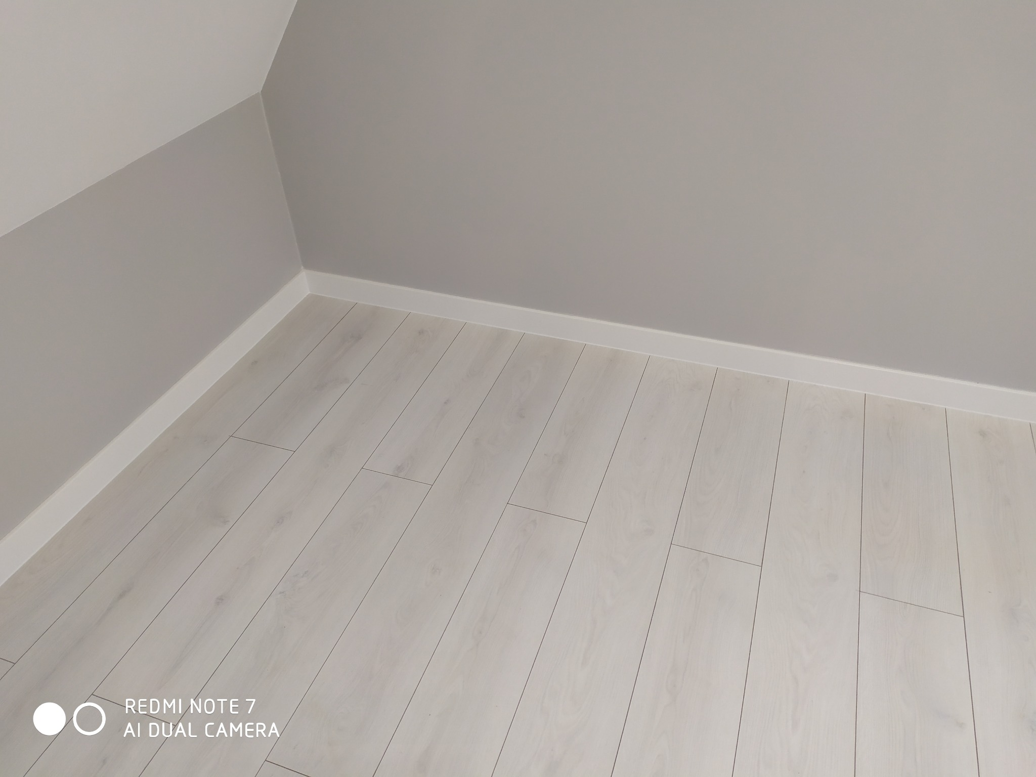 Jasne panele podłogowe ułożone w rogu pokoju z szarą ścianą, widoczne białe listwy przypodłogowe.