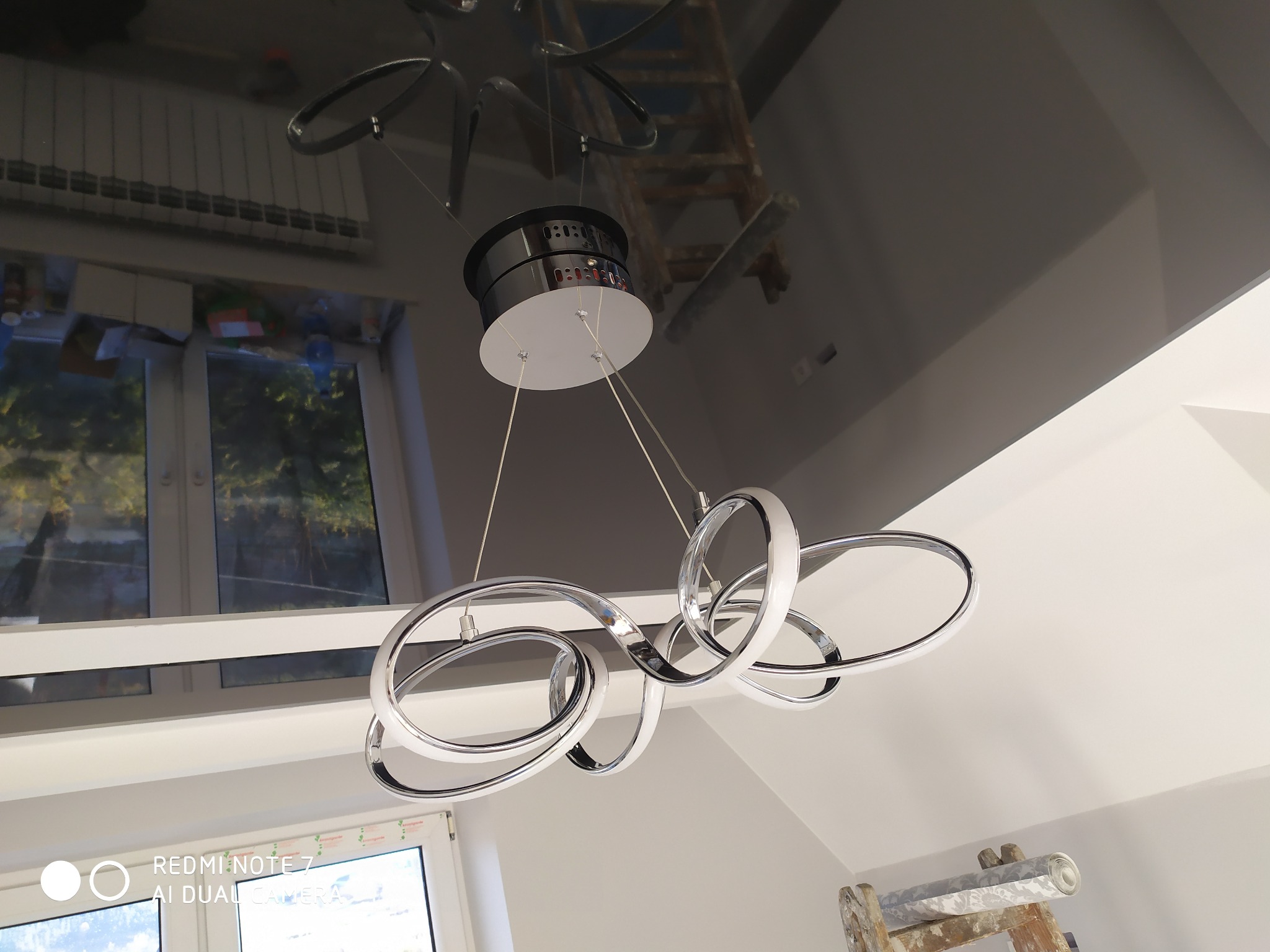 Błyszczący, czarny sufit z nowoczesną lampą wiszącą w kształcie połączonych okręgów, widoczna drabina malarska i okno.