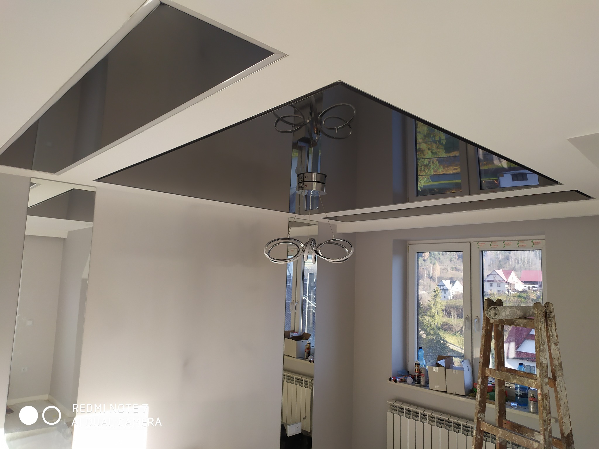 Nowoczesny sufit z podwieszanymi panelami w kolorze grafitowym, odbijający światło lampy, drabina malarska przy oknie, widok na zabudowania za oknem.