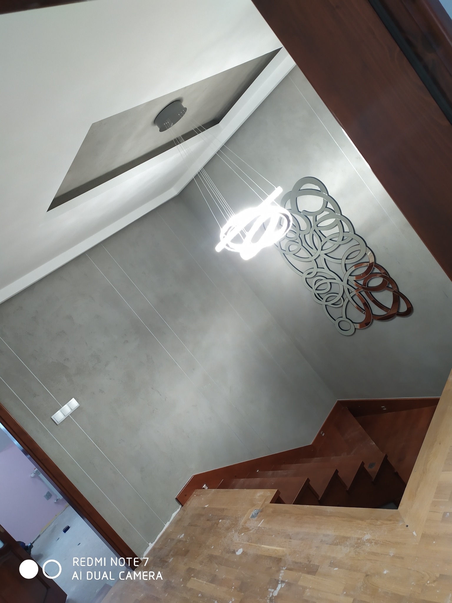 Nowoczesne wnętrze z szarą, gładką ścianą, drewnianymi schodami i designerską lampą wiszącą z metalowymi elementami, widok z góry na klatkę schodową.