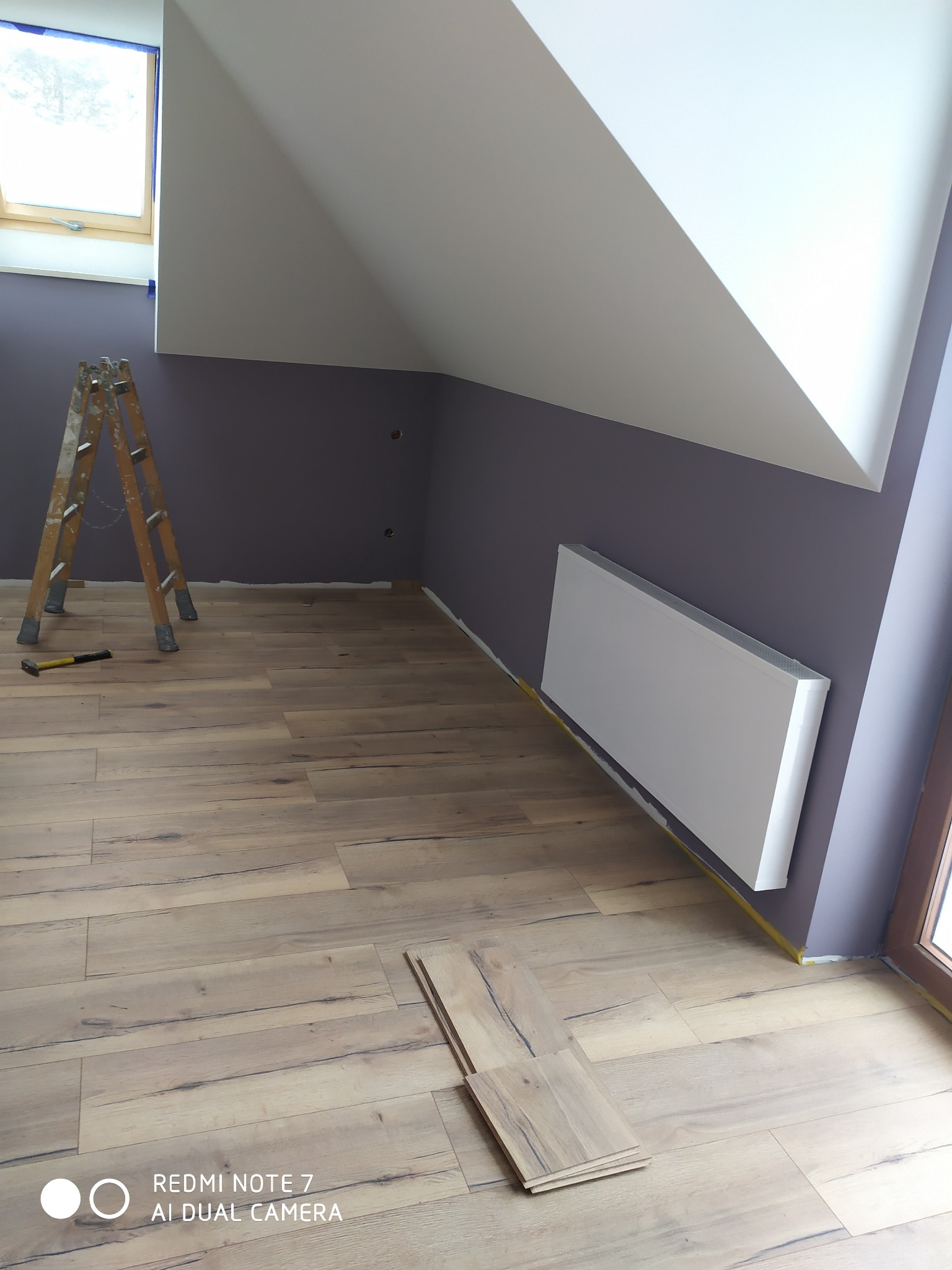 Poddasze po malowaniu ścian na fioletowo, widoczne ułożone panele podłogowe, drabina malarska, grzejnik panelowy i okno dachowe z taśmą malarską.