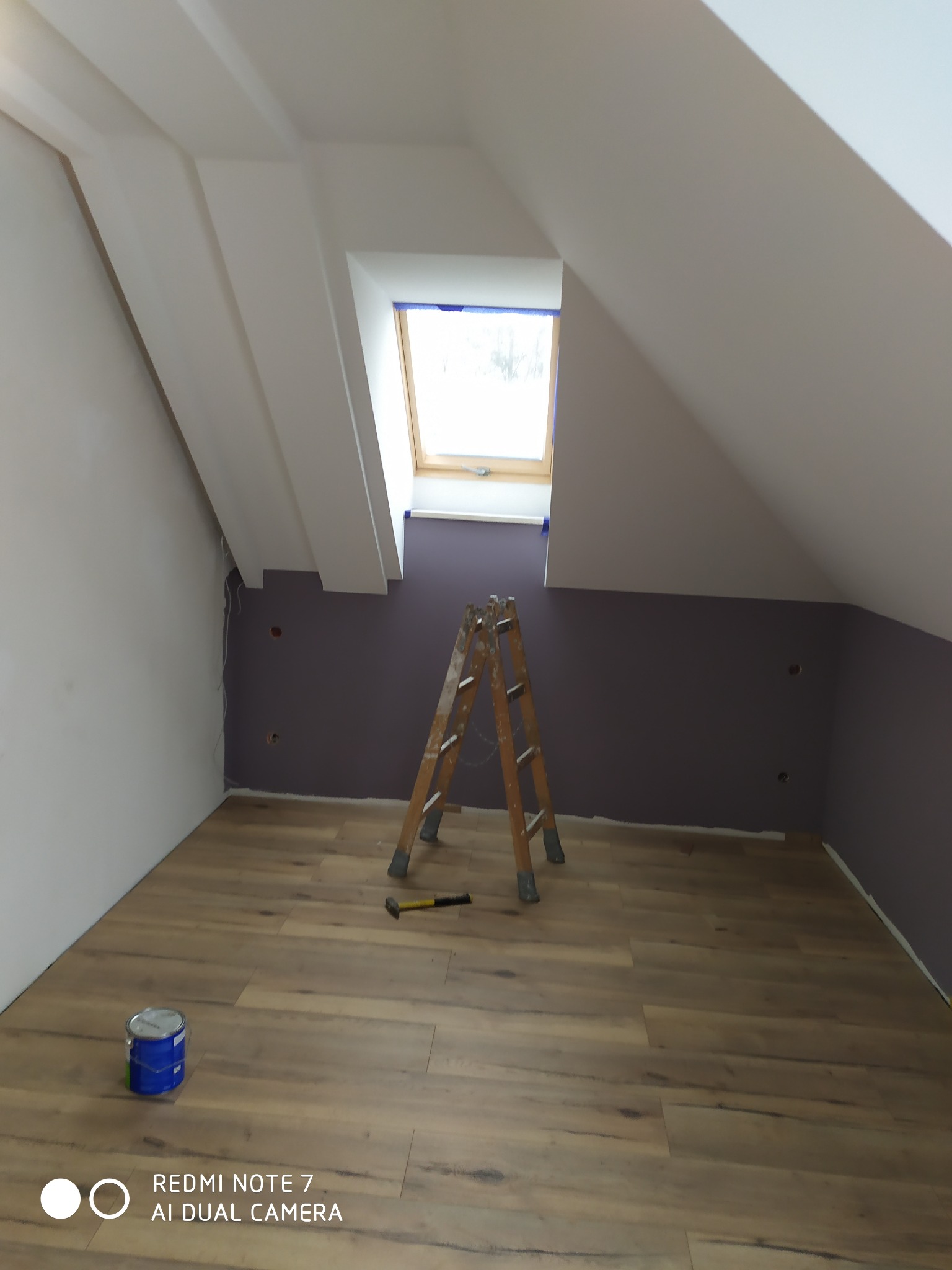 Poddasze w trakcie odświeżania: częściowo pomalowana ściana w kolorze śliwkowym, drewniana drabina, wiadro z farbą i młotek na nowo ułożonych panelach podłogowych.