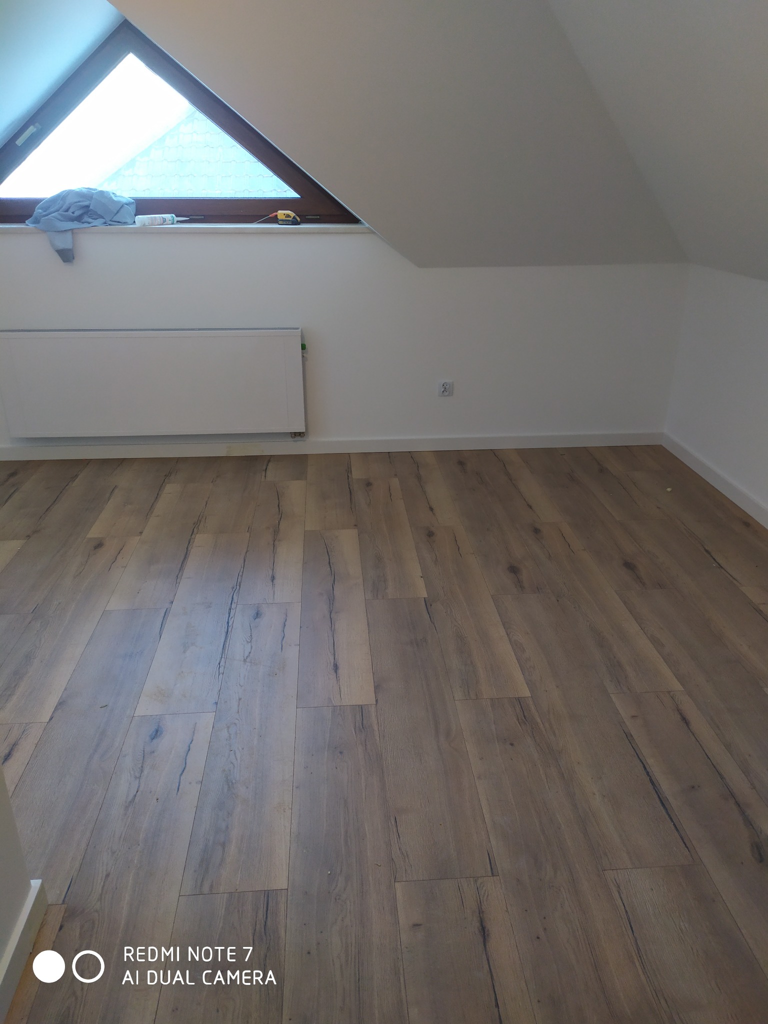 Pokój na poddaszu po remoncie z ułożonymi panelami podłogowymi w odcieniach brązu, widoczne okno trójkątne, białe ściany i grzejnik.