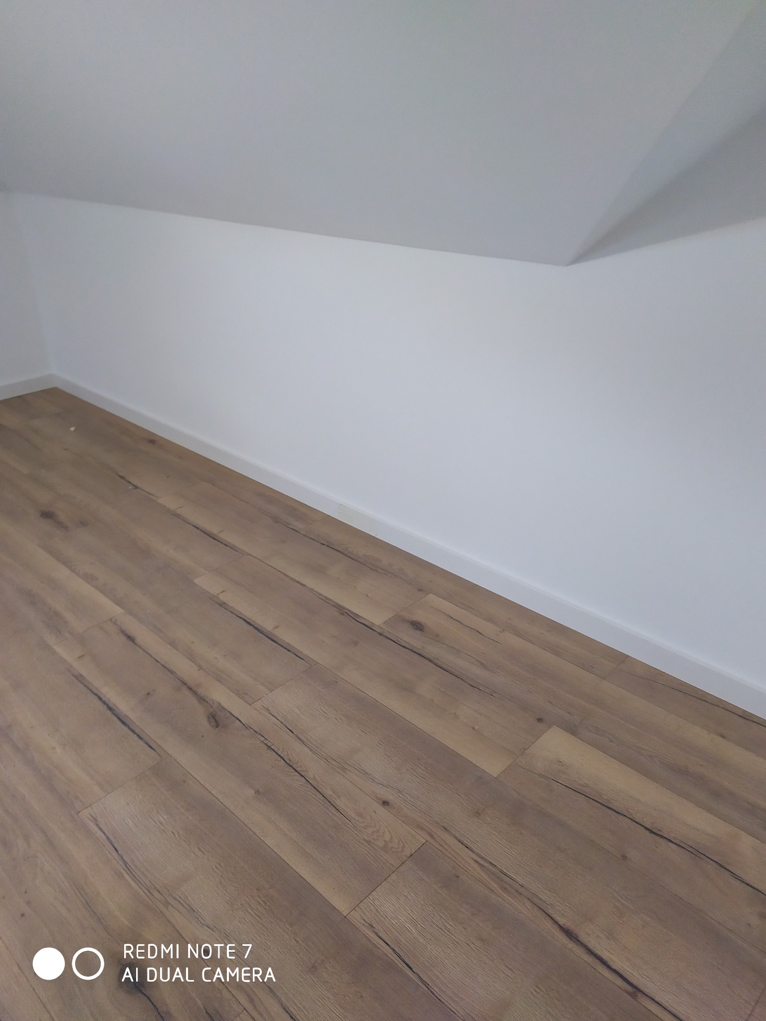 Ukończone wnętrze poddasza ze świeżo ułożonymi panelami podłogowymi o naturalnym, drewnianym wzorze i białymi ścianami z listwami przypodłogowymi.