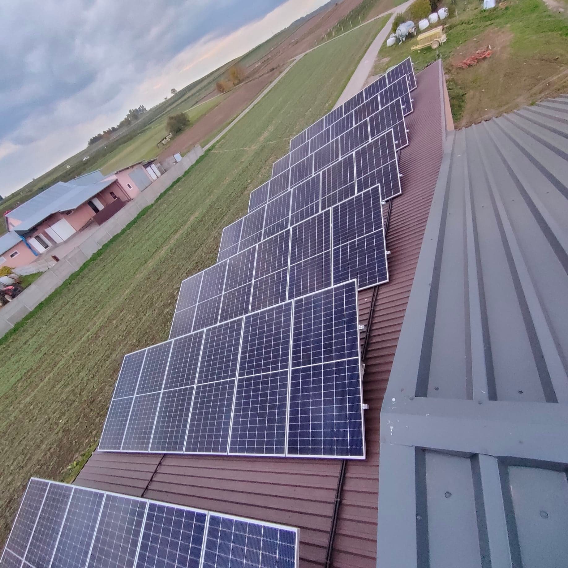 Rząd paneli fotowoltaicznych ułożonych schodkowo na brązowym dachu budynku gospodarczego, z polem uprawnym i zabudowaniami w tle, widok z góry pod kątem.