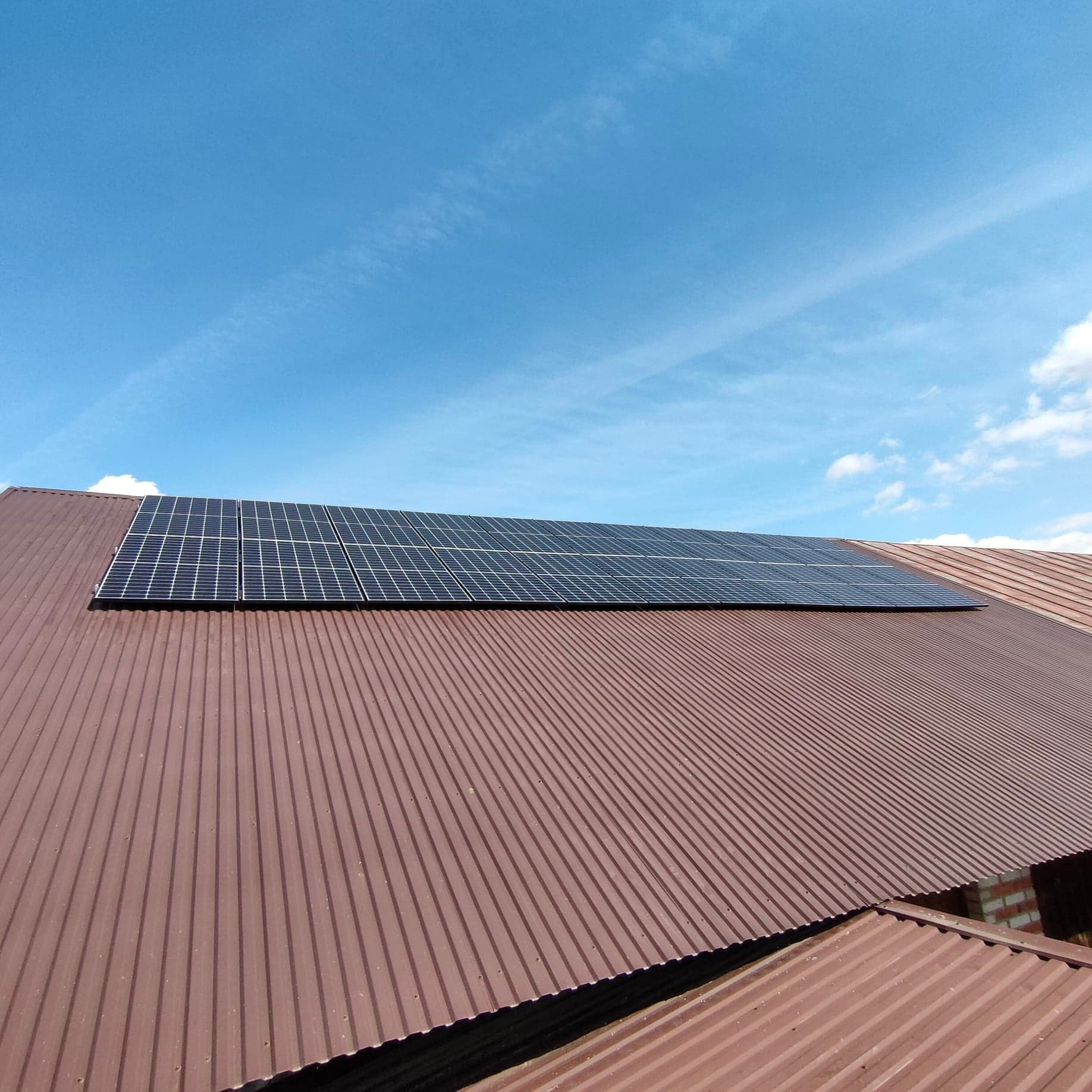 Rząd paneli słonecznych zamontowany na brązowym, falistym dachu pod błękitnym niebem z delikatnymi chmurami.