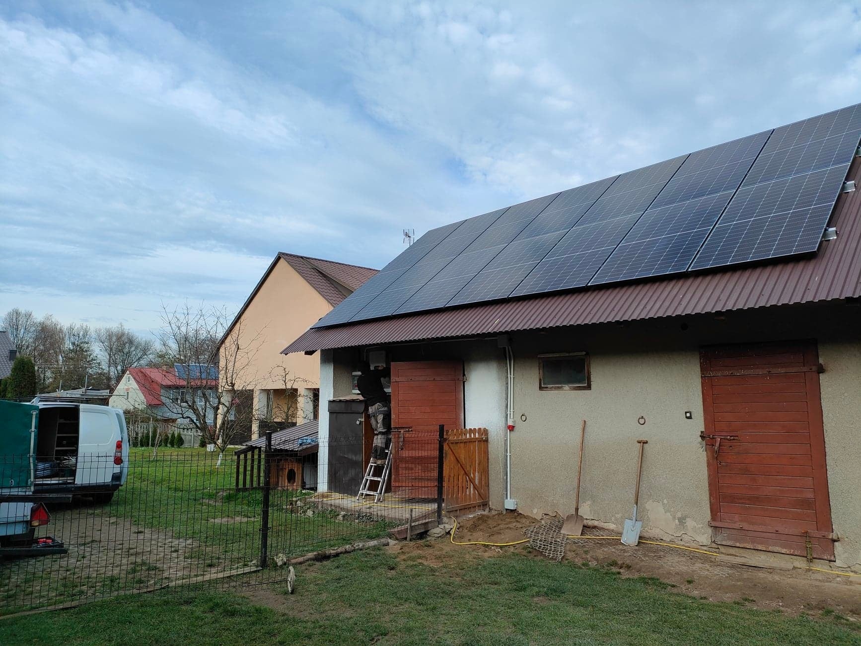 Panele słoneczne zamontowane na dachu budynku gospodarczego z blachy falistej; widoczny pracownik na drabinie, instalujący okablowanie; w tle dom mieszkalny i samochód dostawczy za ogrodzeniem.