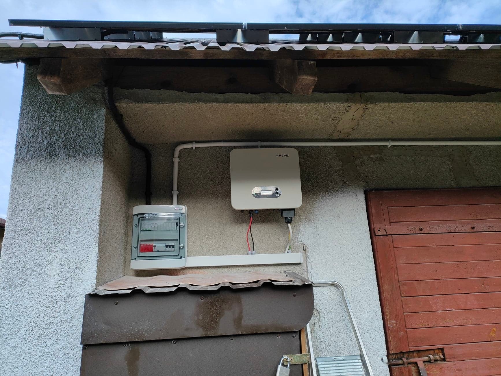 Zamontowany inwerter SOFAR obok skrzynki elektrycznej na elewacji budynku z widocznymi przewodami i elementami instalacji fotowoltaicznej na dachu.