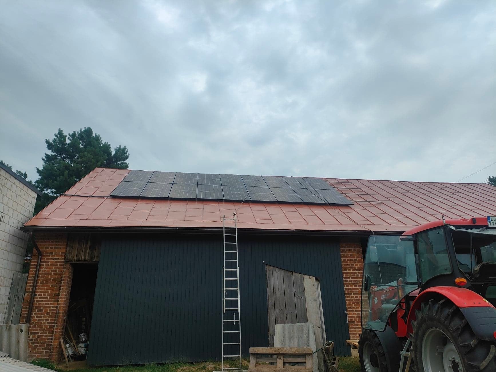 Panele fotowoltaiczne zainstalowane na dachu stodoły z czerwonej dachówki, drabina oparta o ścianę, w tle widoczny czerwony traktor i pochmurne niebo.