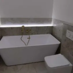 Biała, prostokątna wanna z złotą armaturą i podświetleniem LED, obok podwieszana toaleta w łazience wykończonej marmurem w Sławie.