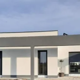 Nowoczesny, parterowy dom z płaskim dachem, świeżo po malowaniu elewacji na biało z szarymi akcentami, z częściowo zasłoniętymi roletami okiennymi.