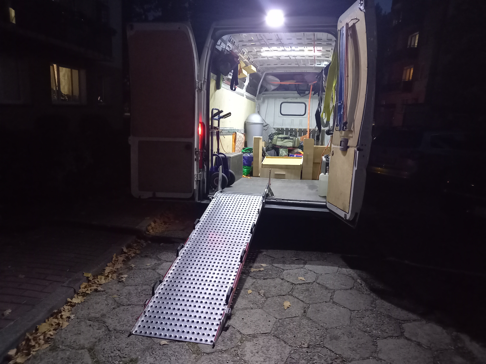 Widok z tyłu otwartego busa z rozłożoną aluminiową rampą, ułatwiającą załadunek, wnętrze częściowo załadowane różnymi przedmiotami, widoczne oświetlenie wewnętrzne.