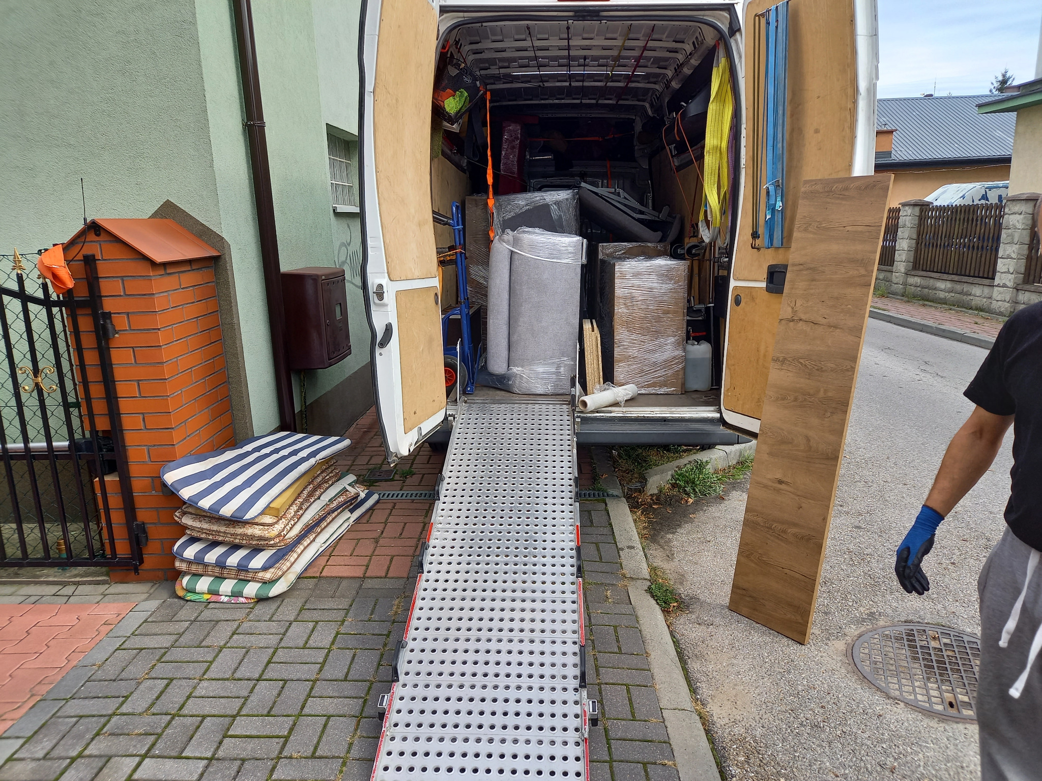 Otwarty furgon z rampą załadunkową, wypełniony meblami i kartonami zabezpieczonymi folią stretch, obok stos materacy na chodniku i pracownik w szarych spodniach dresowych trzymający płytę meblową.