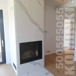 Nowoczesny kominek obudowany płytami imitującymi biały marmur z szarymi żyłami, z czarnym wkładem kominkowym, na podłodze panele imitujące jasne drewno.