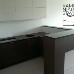 Nowoczesna kuchnia z ciemnym blatem i drewnopodobnymi frontami szafek, logo firmy kamieniarskiej na ścianie.