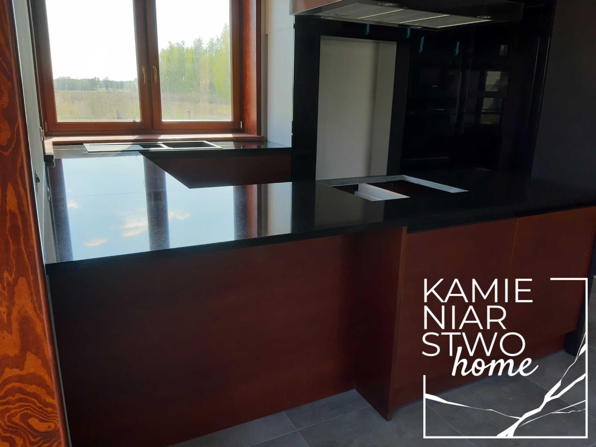 Czarny blat w kuchni z widocznym oknem i logo firmy Kamieniarstwo Home w prawym dolnym rogu.