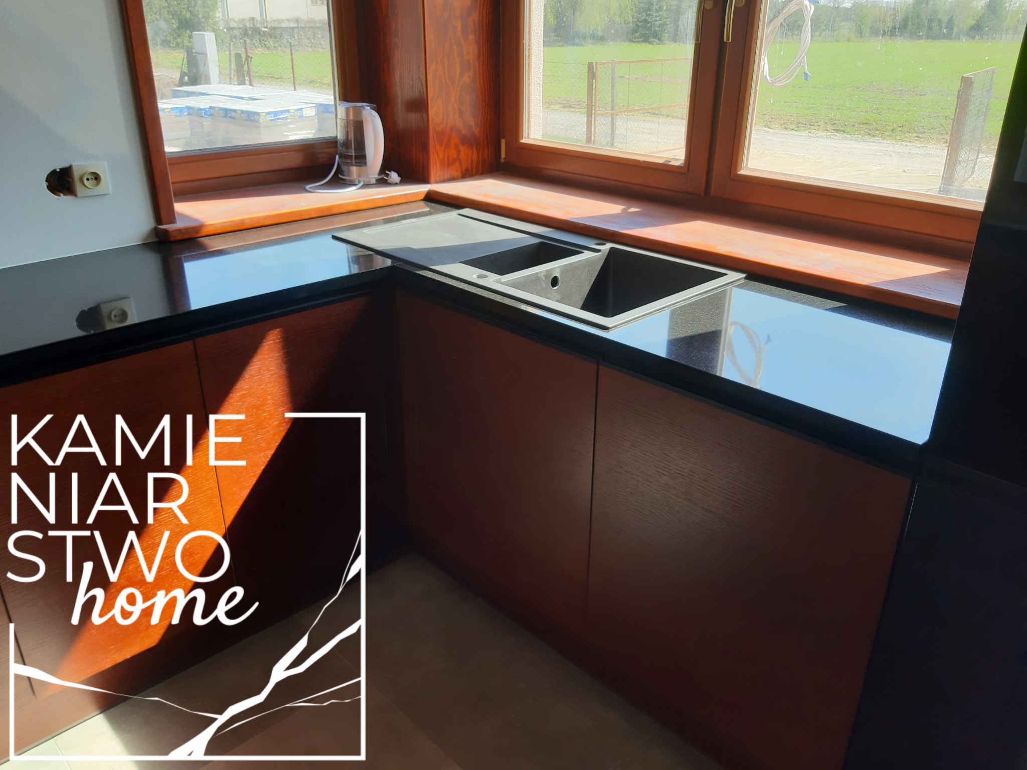 Czarny blat z kamienia z wbudowanym zlewozmywakiem w kuchni w Mińsku Mazowieckim, widoczne drewniane szafki i okno z widokiem na zewnątrz. W lewym dolnym rogu logo firmy kamieniarskiej.