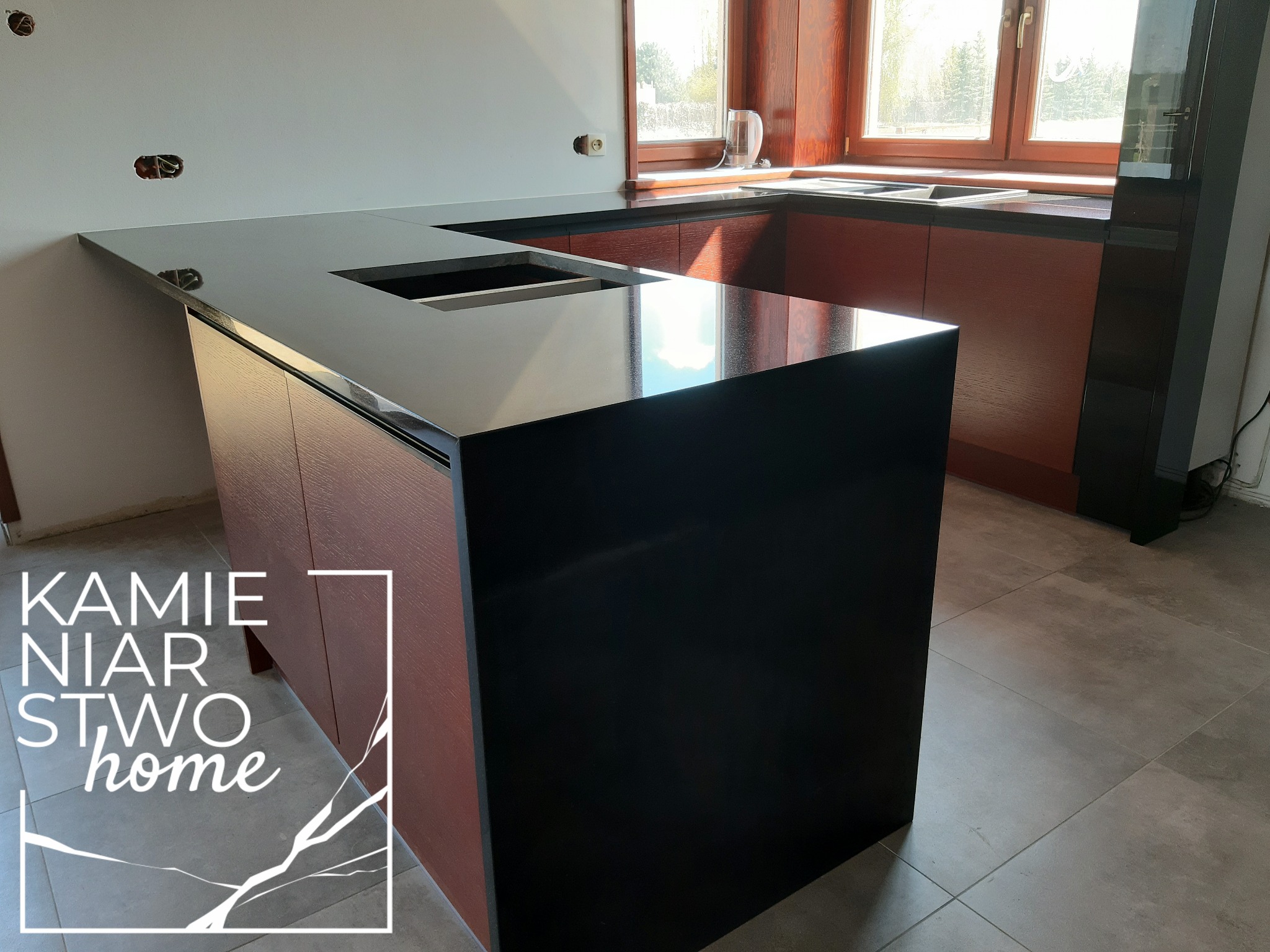 Częściowo zamontowany, czarny blat z otworem na zlew w nowoczesnej kuchni z drewnianymi szafkami i oknem w tle. Widoczne niedokończone prace elektryczne na ścianie.