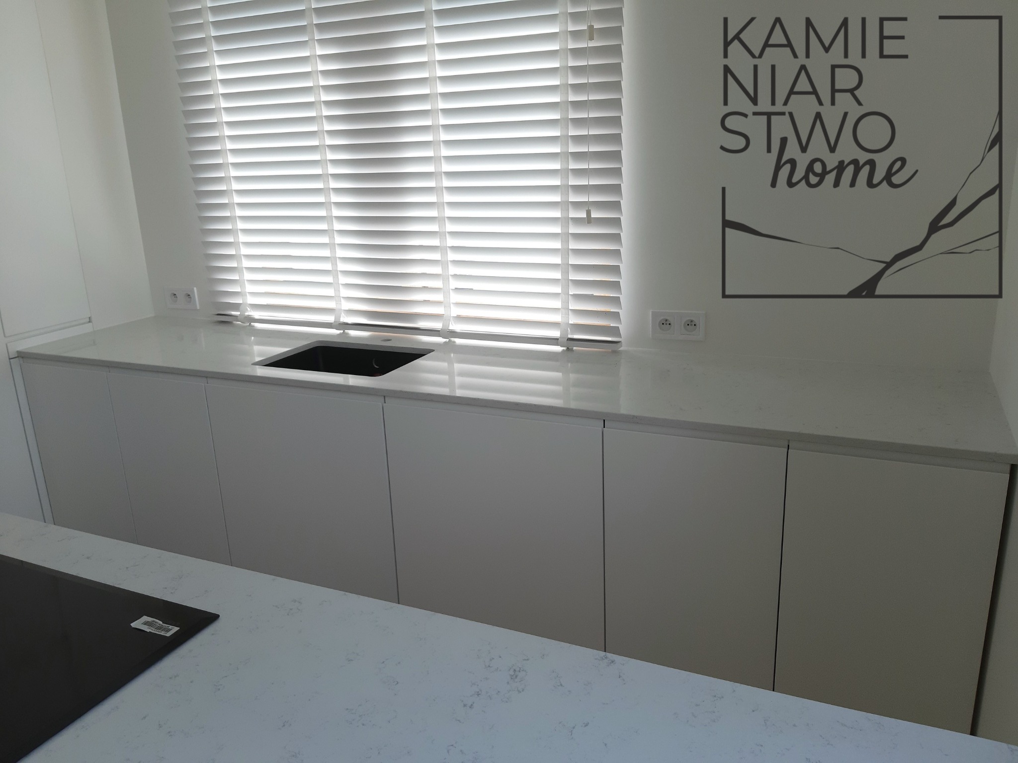 Jasny blat kamienny z konglomeratu Santamargherita Lyskamm z wbudowanym czarnym zlewem, białe szafki kuchenne pod spodem i żaluzje w tle, logo firmy na ścianie.