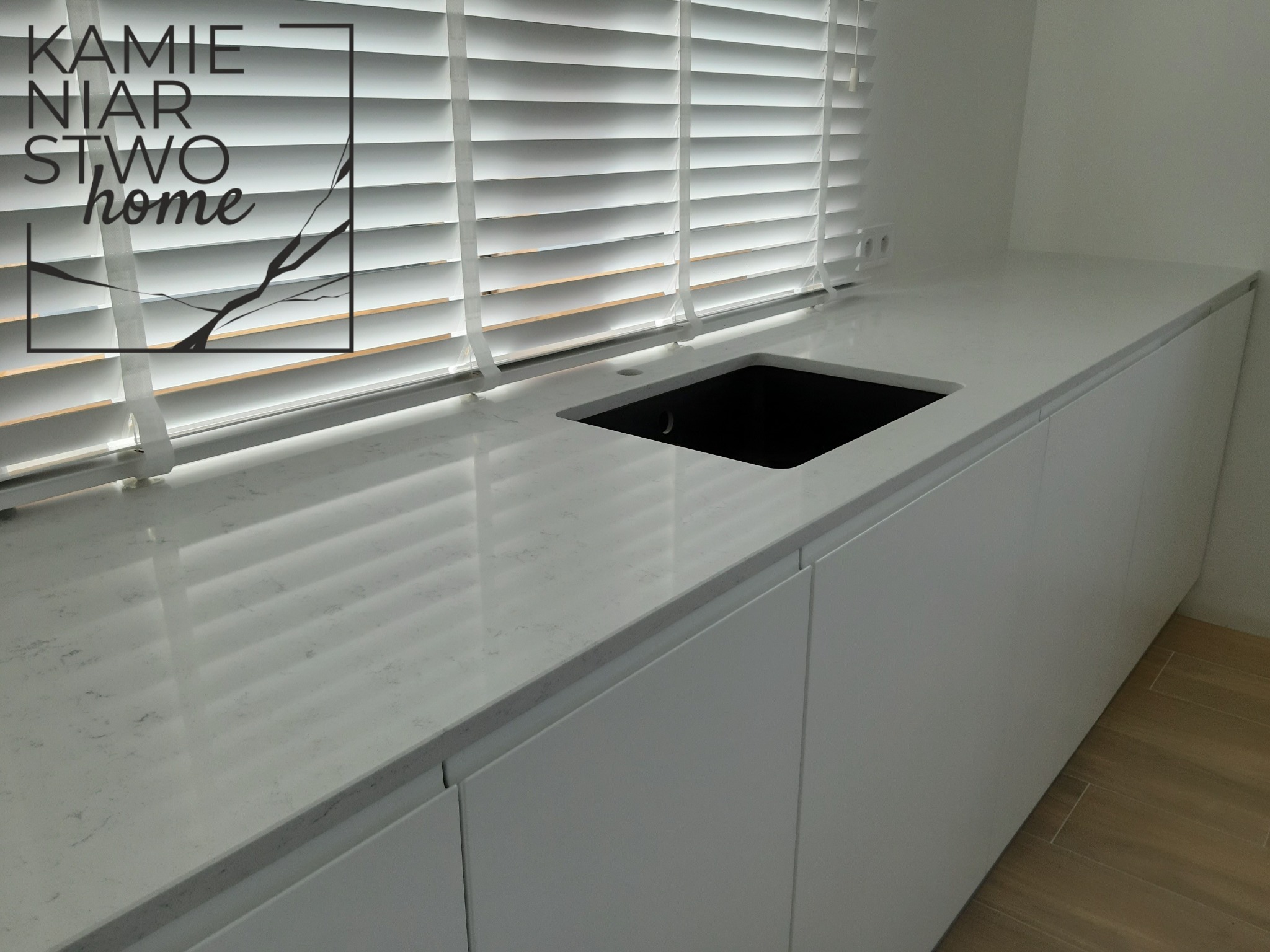 Jasny blat kuchenny z konglomeratu Santamargherita Lyskamm, z wbudowanym czarnym zlewem, oświetlony przez żaluzje okienne.