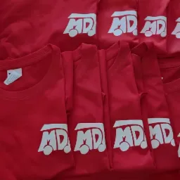 Stos czerwonych koszulek z białym nadrukiem logo w kształcie autobusu z napisem MD, leżące jedna na drugiej.