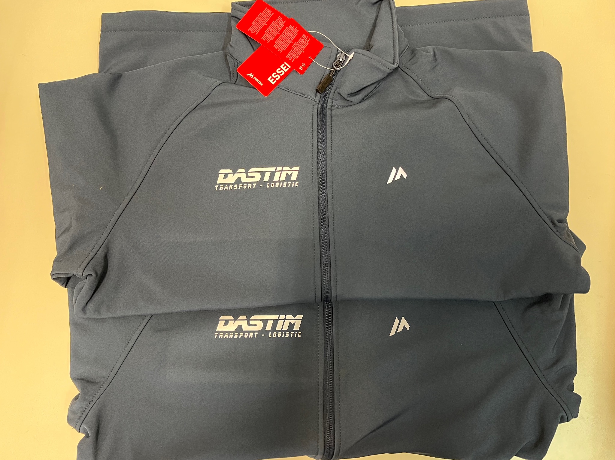 Granatowa kurtka softshell z nadrukowanym logo firmy transportowej 'DASTIM' i czerwoną metką 'ESSEI'.