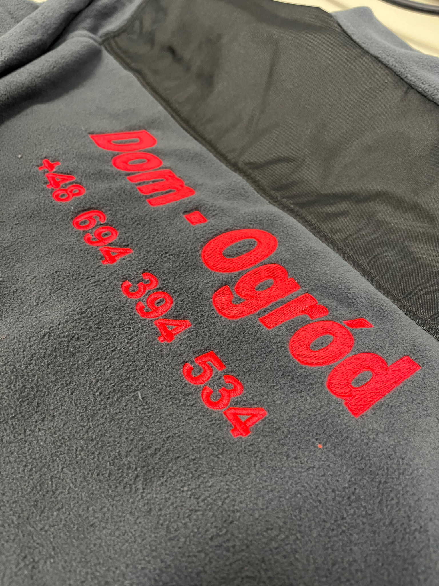 Szara bluza polarowa z czerwonym haftowanym napisem 'Dom-Ogród' oraz numerem telefonu, widoczny fragment czarnego panelu na ramieniu.