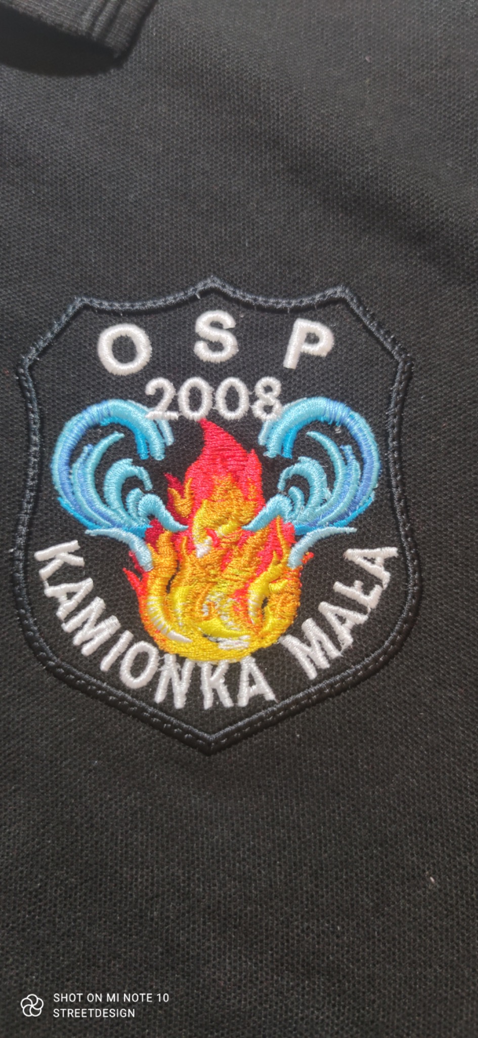 Czarna koszulka polo z wyhaftowanym emblematem OSP Kamionka Mała z motywem płomieni i wody, rok założenia 2008.