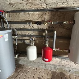 SUNKO GRZEGORZ MICHAŁOWSKI, SZYMON PIEKUTOWSKI S.C. - Dwa zbiorniki buforowe Sunko i Galmet w kotłowni w Białymstoku, z widoczną instalacją hydrauliczną i manometrami na tle surowej ściany.