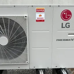 Zewnętrzna jednostka pompy ciepła LG Therma V z widocznymi naklejkami firmy Sunko, zamontowana na zewnątrz budynku, pokryta kroplami deszczu.