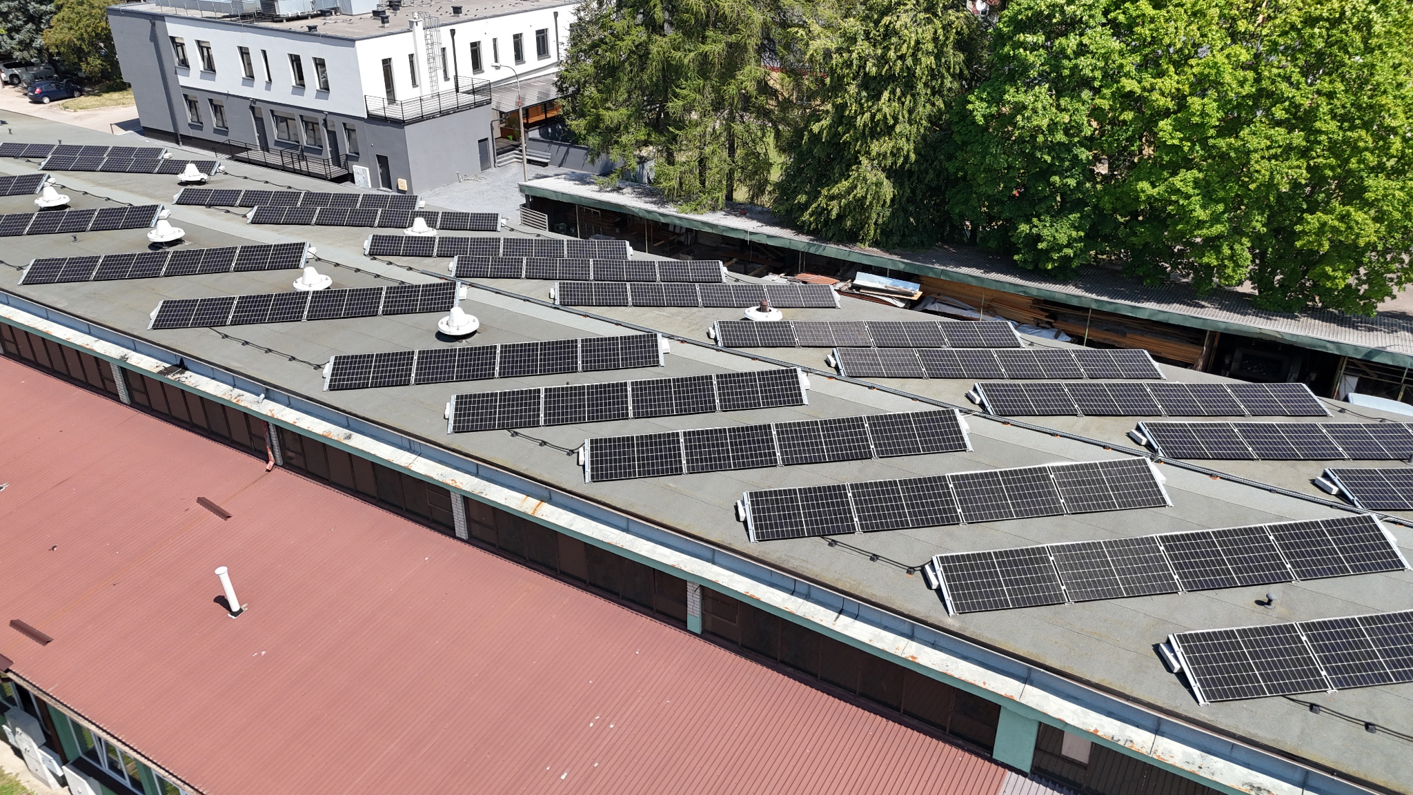 Widok z góry na panele słoneczne zamontowane rzędami na płaskim dachu budynku przemysłowego, z widocznymi elementami wentylacji i fragmentami sąsiednich budynków oraz zieleni.