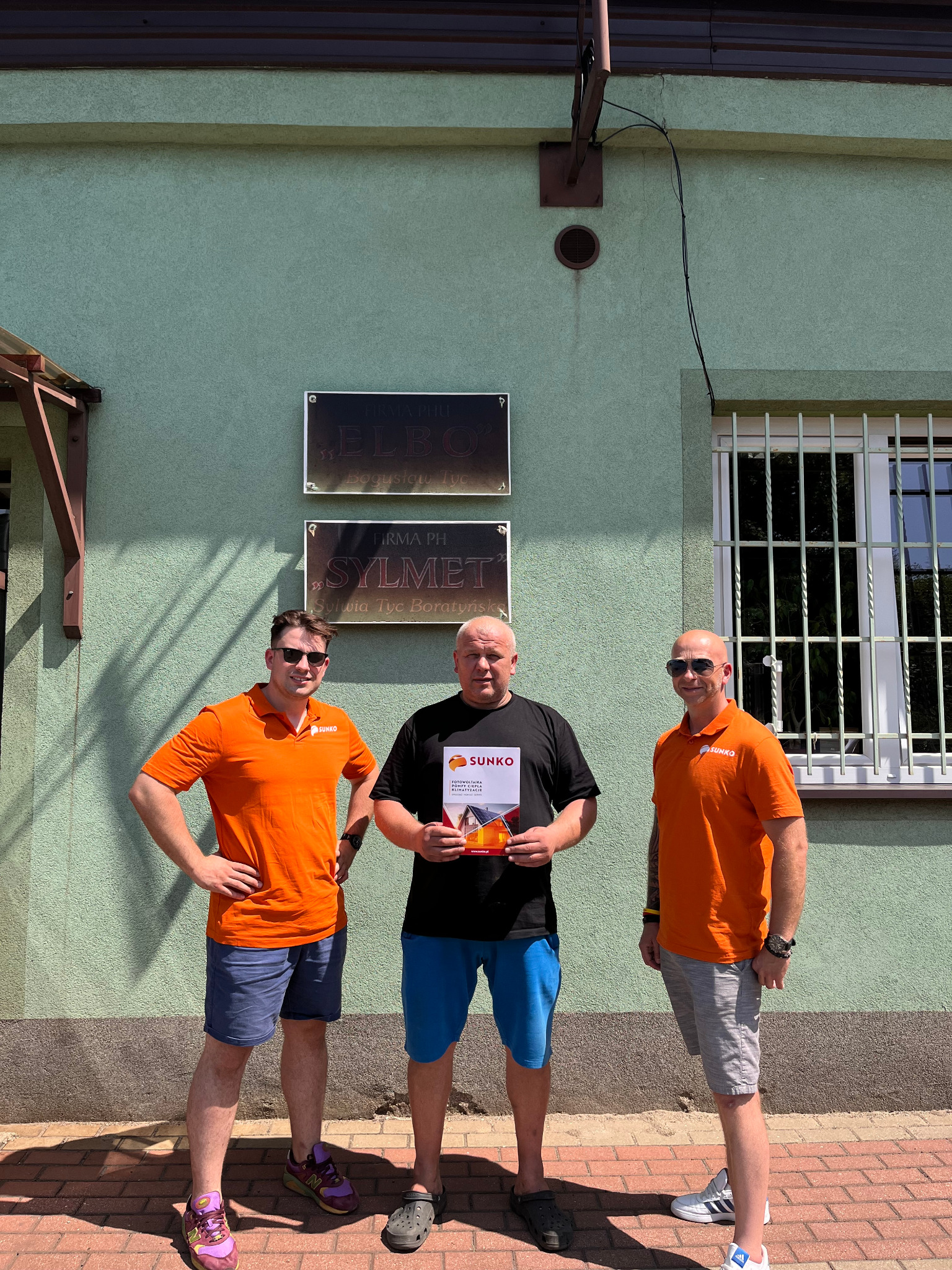 Trzech mężczyzn w pomarańczowych koszulkach z logo firmy SUNKO stoi przed budynkiem z tabliczkami informacyjnymi firm ELBO i SYLMET, środkowy mężczyzna trzyma folder reklamowy SUNKO z informacjami...