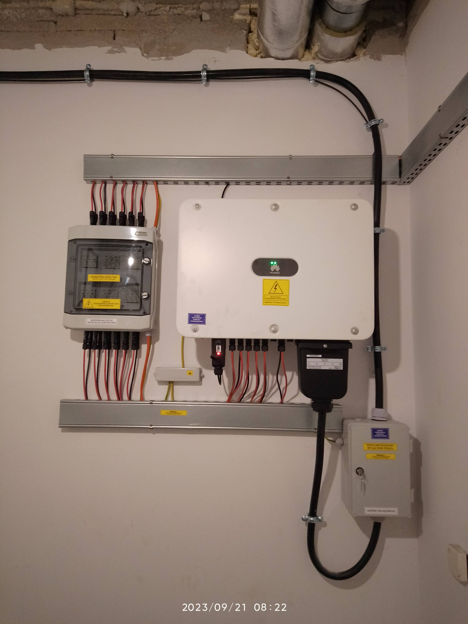 Rozdzielnica PV DC i inwerter Huawei zamontowane na ścianie wewnątrz budynku, widoczne przewody i zabezpieczenia elektryczne.