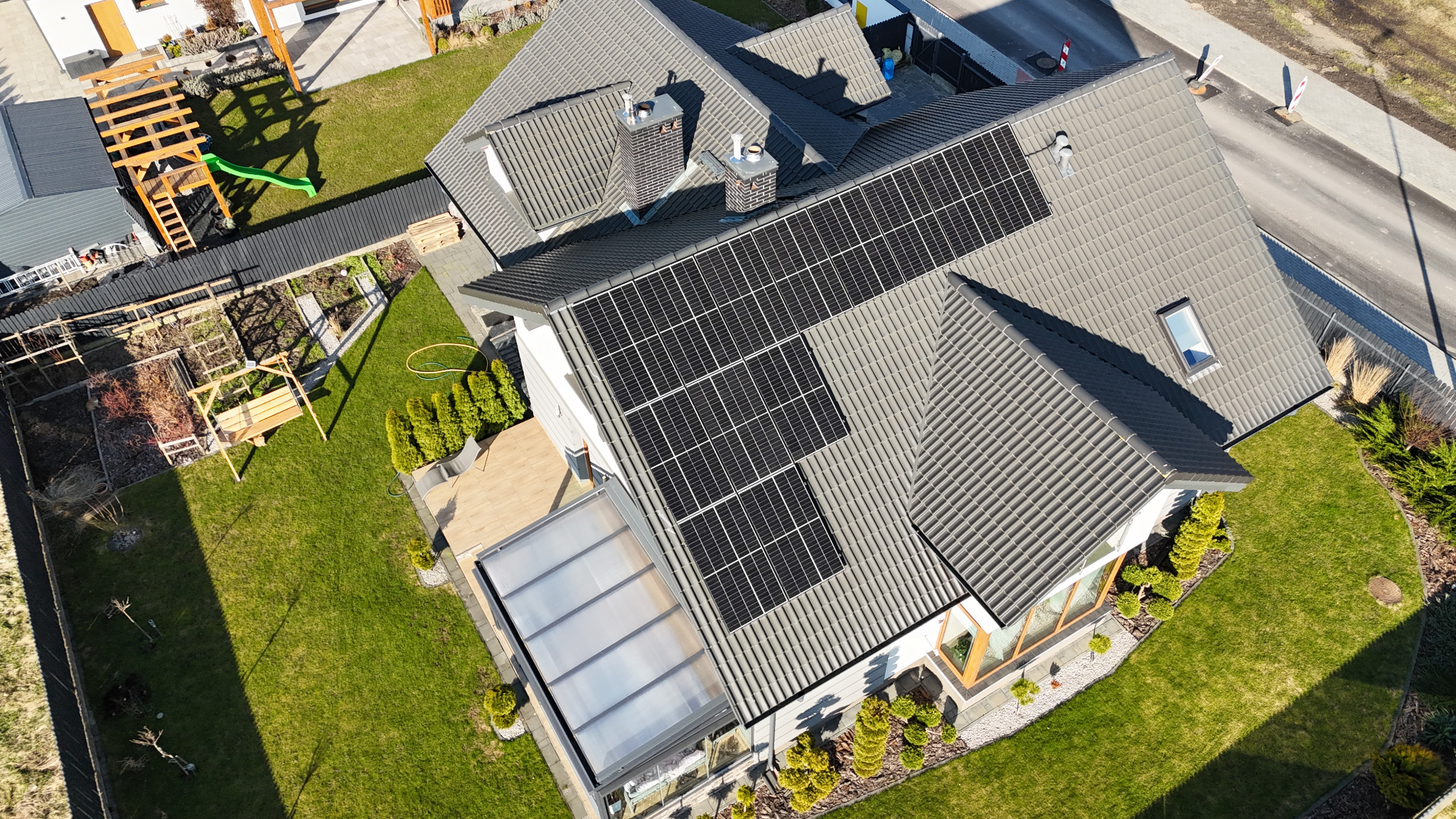 Widok z lotu ptaka na dom jednorodzinny z szarą dachówką i panelami słonecznymi na dachu, zadbany trawnik, plac zabaw w ogrodzie.