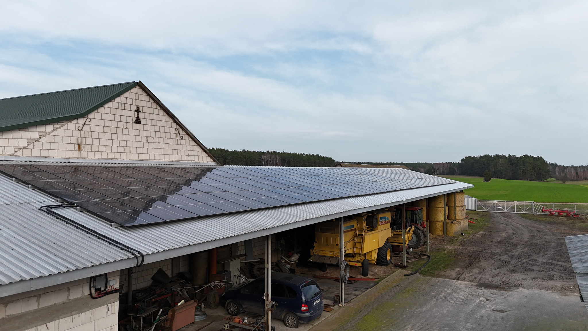 Instalacja fotowoltaiczna na dachu budynku gospodarczego, widoczny kombajn rolniczy i samochód osobowy wewnątrz, tło stanowi pole i las.