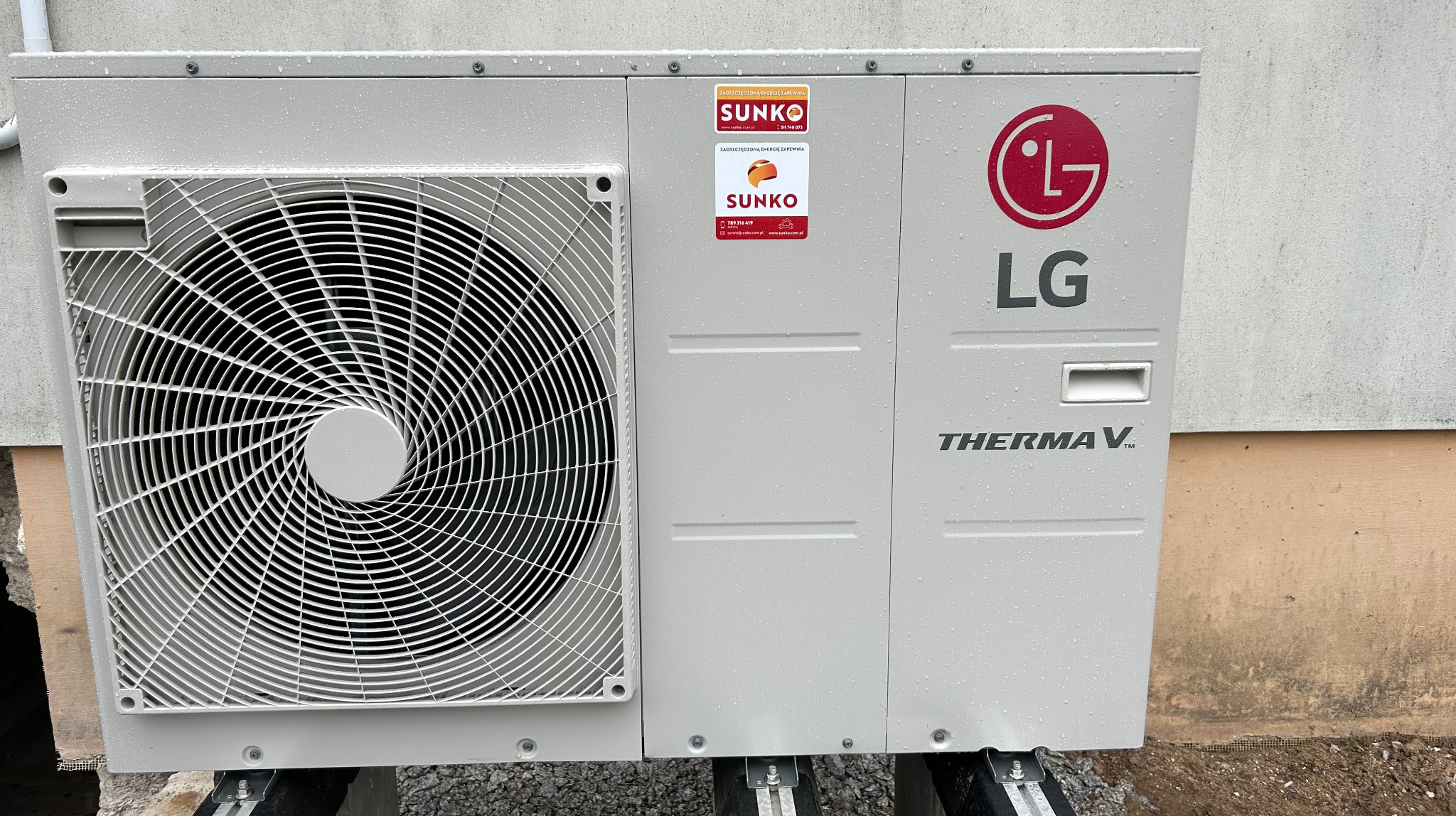 Zewnętrzna jednostka pompy ciepła LG Therma V z widocznymi naklejkami firmy Sunko, zamontowana na zewnątrz budynku, pokryta kroplami deszczu.