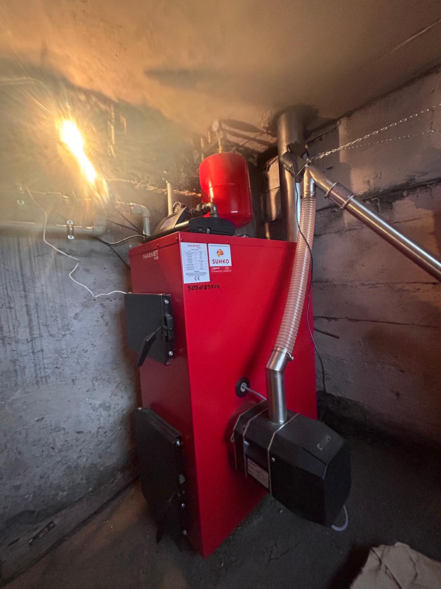Czerwony piec Narmet Plus z podajnikiem i zasobnikiem, zainstalowany w kotłowni z betonowymi ścianami, oświetlony górnym światłem.