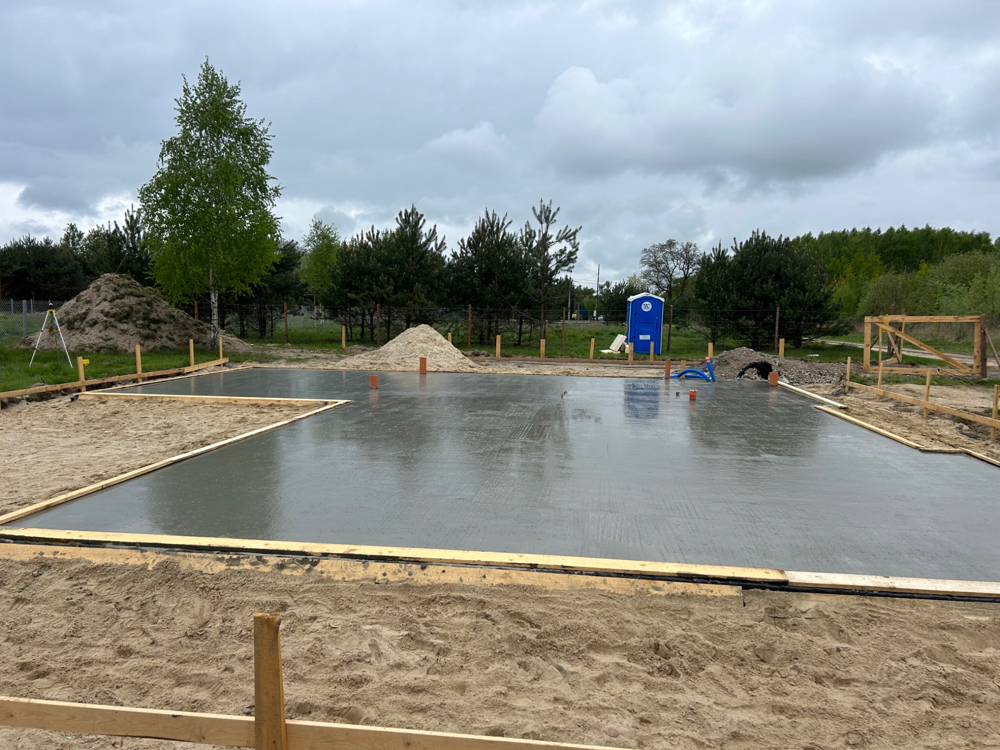 Świeżo wylana, mokra płyta fundamentowa otoczona drewnianym szalunkiem na piaszczystym podłożu, widoczny stos piasku, drzewa i przenośna toaleta w tle, pochmurne niebo.