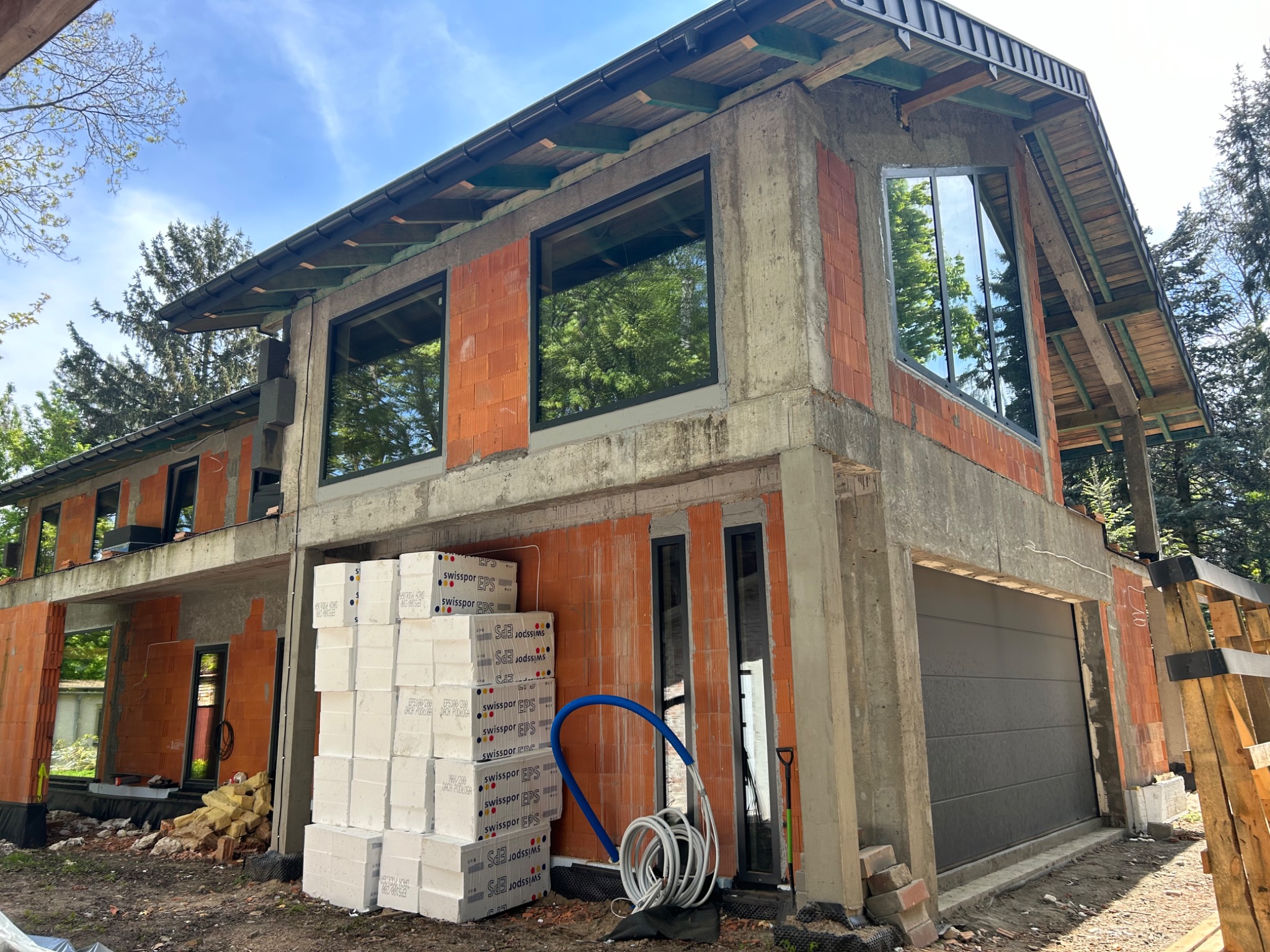 Surowy stan budynku żelbetowego z widocznymi elementami konstrukcyjnymi, pustakami ceramicznymi i oknami w czarnych ramach, z ułożonymi obok paczkami styropianu Swissopor EPS i niebieskim wężem...